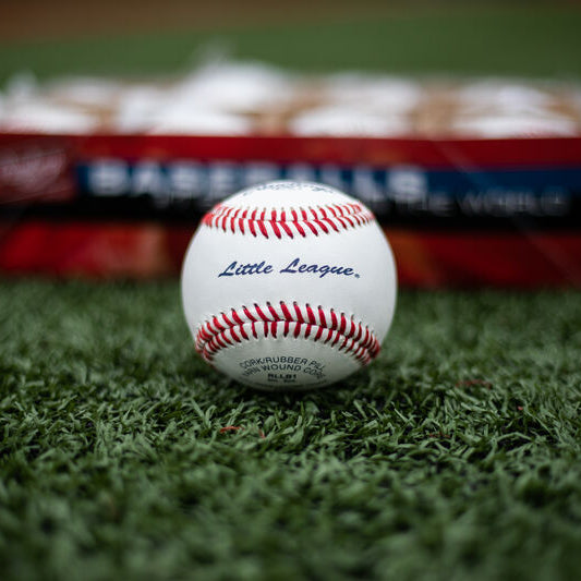 A Rawlings Little League baseball with red stitching sits centered on green turf in front of a blurred baseball box.
