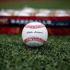 A Rawlings Little League baseball with red stitching sits centered on green turf in front of a blurred baseball box.